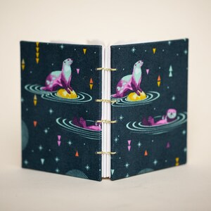 Puede incluir: Diario abierto con una cubierta azul oscuro que presenta un patr&oacute;n repetido de nutrias en una escena acu&aacute;tica. Las nutrias son rosas y turquesas, con encuadernaci&oacute;n dorada y p&aacute;ginas blancas. El dise&ntilde;o incluye estrellas y formas geom&eacute;tricas.