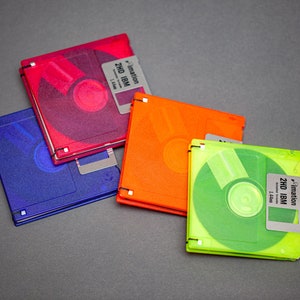 Pu&ograve; includere: Quattro floppy disk da 3,5 pollici colorati in rosso, blu, arancione e verde. Ogni disco ha un'etichetta con il testo "imation 2HD IBM 1.44MB". I dischi sono disposti su una superficie grigia.