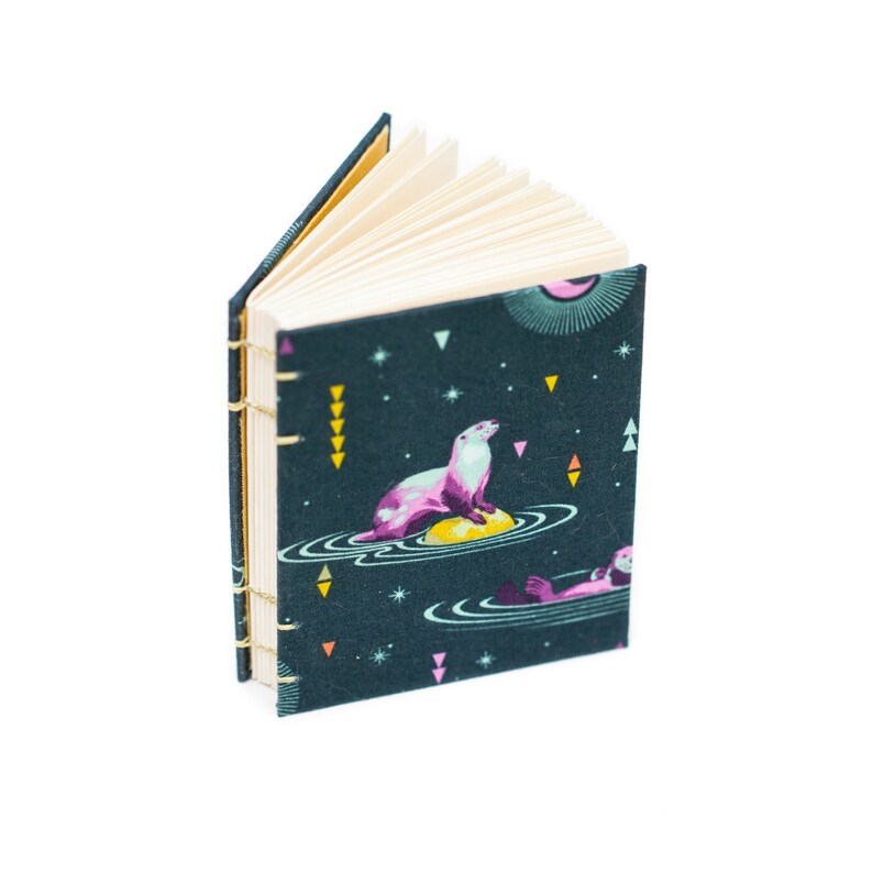 Puede incluir: Un peque&ntilde;o cuaderno abierto con una cubierta de color verde azulado oscuro que presenta un dise&ntilde;o caprichoso de nutria. La nutria est&aacute; representada en tonos de rosa y morado, flotando sobre una esfera amarilla. El cuaderno est&aacute; encuadernado con hilo dorado y tiene p&aacute;ginas color crema.