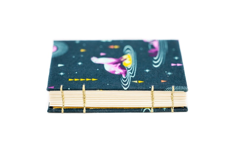 Puede incluir: Un peque&ntilde;o cuaderno rectangular con una cubierta azul oscuro que presenta un patr&oacute;n de tem&aacute;tica espacial con planetas y estrellas. Las p&aacute;ginas son de color crema y la encuadernaci&oacute;n est&aacute; asegurada con hilo dorado.