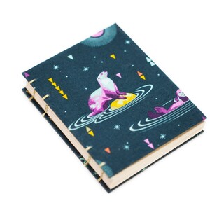 Puede incluir: Un peque&ntilde;o cuaderno de tapa dura con una cubierta azul oscuro que presenta un dise&ntilde;o caprichoso con tem&aacute;tica espacial. La cubierta representa una nutria marina rosa y blanca sobre un planeta amarillo, rodeada de estrellas y planetas. El cuaderno est&aacute; encuadernado con hilo dorado.