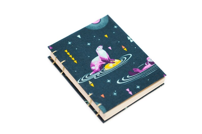Puede incluir: Un peque&ntilde;o cuaderno de tapa dura con una cubierta azul oscuro que presenta un dise&ntilde;o caprichoso con tem&aacute;tica espacial. La cubierta representa una nutria marina rosa y blanca sobre un planeta amarillo, rodeada de estrellas y planetas. El cuaderno est&aacute; encuadernado con hilo dorado.