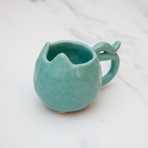 May include: A turquoise ceramic mug with a unique, rounded shape and a handle. The rim of the mug has a wavy design. The handle has a decorative fin-like detail. The mug is a solid color and appears to be handmade.