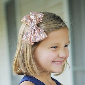 Puede incluir: Una niña pequeña con el pelo rubio corto que lleva un top azul sin mangas y unos vaqueros azules. Lleva un lazo rosa brillante en el pelo. Está sonriendo y mirando hacia un lado.