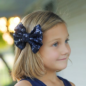 May include: A young girl with blonde hair wearing a navy blue tank top and a sparkly blue bow in her hair. She is standing in front of a white porch.