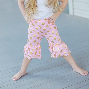 Puede incluir: Una niña pequeña con un pantalón de volantes de lunares rosa y dorado y una diadema de flores rosa. Está de pie en un porche de madera.