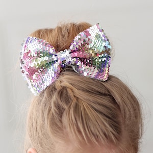 May include: A young girl with blonde hair wearing a sparkly silver top and a large iridescent sequin bow in her hair.