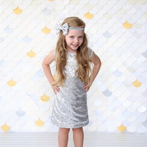 Puede incluir: Una niña con un vestido plateado de lentejuelas sin mangas y una diadema a juego con un lazo. El vestido es recto y llega a las rodillas. El fondo presenta un patrón de escamas blanco, dorado y azul claro.