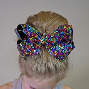May include: A young girl with blonde hair wearing a grey tank top and a large, colorful sequined bow in her hair.