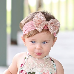 May include: A baby girl wearing a white floral dress with a lace neckline and a pink headband with a gold sequin bow.