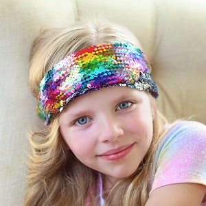May include: A young girl with long blonde hair wearing a rainbow sequin headband and a tie-dye shirt.