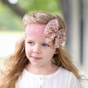 May include: A young girl with long blonde hair wearing a white dress with gold accents. She is wearing a pink lace headband with a large gold sequin bow.
