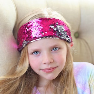 May include: A young girl with long blonde hair wearing a pink sequined headband. She is wearing a rainbow tie-dye shirt.
