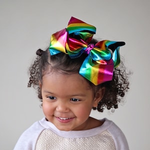 May include: A young girl with curly brown hair is wearing a white and gold sweater with a rainbow iridescent bow in her hair.