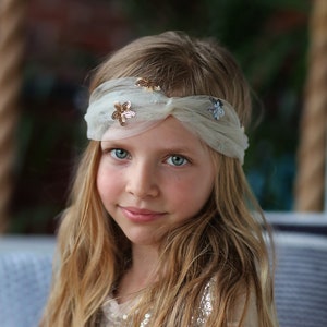 May include: A young girl with long blonde hair wearing a white headband with gold and silver sequins. The headband is tied in a knot at the top of her head.