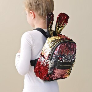 Puede incluir: Una mochila de lentejuelas rojas, doradas y rosas con detalles negros y correas de orejas de conejo.