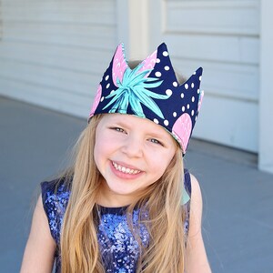 May include: A young girl wearing a blue sequined top and a crown with a pineapple and polka dot pattern.