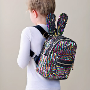 May include: A multi-coloured sequin backpack with black faux leather trim and bunny ear accents. The backpack has a zipper closure and a small front pocket.