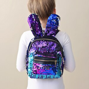 May include: A purple, blue, and pink sequined backpack with bunny ears. The backpack has a black zipper and a black leather patch on the front.
