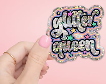 May include: A holographic sticker with the words "Glitter Queen" in a black, cursive font. The sticker has a rainbow border and colorful star accents. The background is a solid pink.
