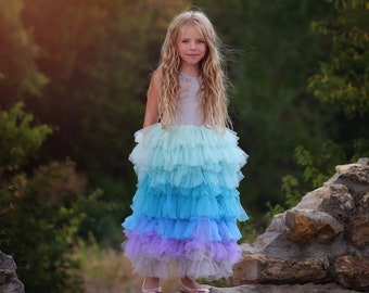 May include: A young girl wearing a light blue, layered tulle dress with a white bodice. She is standing on a stone wall with a green background.