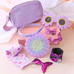 May include: A purple and lavender gift box filled with various items, including a purple headband, a purple and blue pop-it toy, a purple and black sequin wrist cuff, a purple and pink mermaid tail keyring, and two purple butterfly charms.