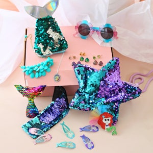 May include: A mermaid-themed gift box containing a teal and green sequin mermaid tail purse, a purple and teal sequin star-shaped purse, a rainbow sequin mermaid tail hair clip, a turquoise necklace, and a set of mermaid-themed hair clips.