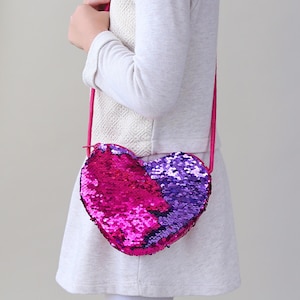 May include: A pink and purple heart-shaped sequin crossbody bag. The bag has a long strap and is made of sparkly sequins.