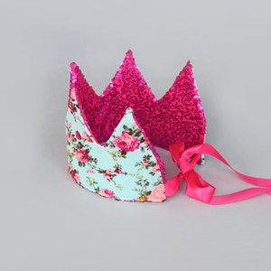 Può includere: Una corona di tessuto rosa e blu con un motivo floreale e paillettes rosa. La corona ha un nastro di raso rosa legato sul retro.