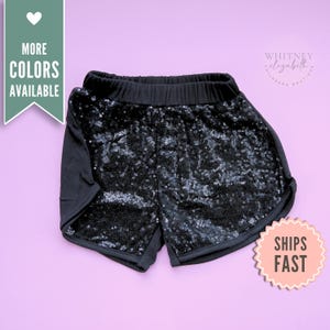 May include: Black sequin shorts with a black elastic waistband. The shorts have a solid black side panel and a rounded hem. The image includes a pink sticker that says "SHIPS FAST" and a green banner that says "MORE COLORS AVAILABLE".