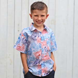 May include: A young boy wearing a white button-down shirt with a blue and orange crab print. He is smiling and has his hands in his pockets. He is wearing navy blue shorts with white polka dots.