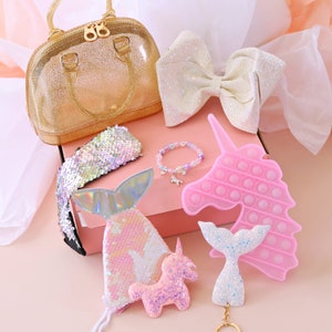 May include: A pink box filled with a variety of items, including a gold glitter purse, a white glitter bow, a pink unicorn pop-it, a mermaid tail keyring, a pink unicorn sequin patch, a mermaid tail sequin patch, and a rainbow sequin headband.