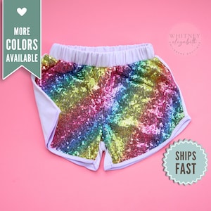 May include: Rainbow sequin shorts with a white trim and elastic waistband. The shorts have a vibrant, multi-colored sequin design. The text "MORE COLORS AVAILABLE" is on the left side of the shorts.