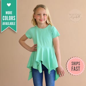 May include: A mint green short-sleeved top with a unique asymmetrical hemline. The top is paired with blue jeans. The image includes text that says "MORE COLORS AVAILABLE" and "SHIPS FAST".