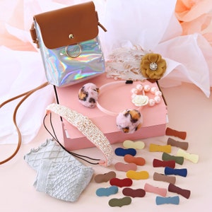 May include: A pink gift box filled with various accessories, including a headband with faux fur pom-poms, a pearl headband, a bracelet with pearls and a flower charm, a pair of socks, and a set of colourful hair clips.