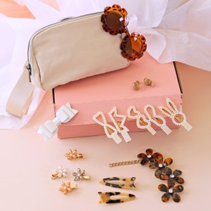 May include: A pink gift box with a white bow, a beige bag, a pair of tortoiseshell sunglasses, and a variety of hair clips, including pearl and flower designs.