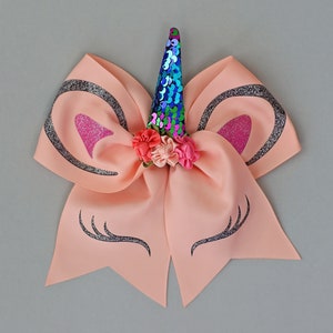 May include: A peach-coloured hair bow with a unicorn design. The bow has glitter accents and a sparkly blue and green horn with pink flowers.