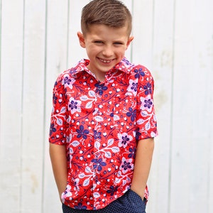 May include: A young boy wearing a red, white, and blue floral print button-down shirt with short sleeves and navy blue pants with white polka dots. He is smiling and has his hands in his pockets.