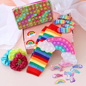 May include: A rainbow striped sock, a rainbow pop-it, and a variety of rainbow hair clips and earrings are displayed in a pink gift box. The box is open and the items are arranged neatly.
