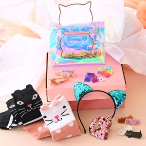 May include: A pink gift box containing a clear iridescent cat-shaped purse, a headband with teal sequin cat ears, black and white cat socks, and pink and white cat socks with hearts. There are also several colourful cat-shaped hair clips.