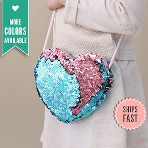 May include: A heart-shaped crossbody bag covered in reversible sequins that transition between light blue and rose gold colors. The bag features a thin, light pink shoulder strap and a zipper closure along the top edge of the heart-shaped frame.