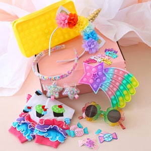 May include: A pink and yellow gift box filled with colourful accessories, including a unicorn headband, a rainbow pop-it, and a pair of rainbow sunglasses. The box also contains a pair of leg warmers with a cupcake design and several colourful hair clips.