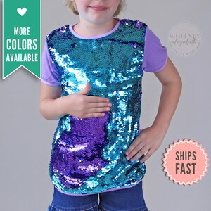 Puede incluir: Una camiseta con lentejuelas reversibles con lentejuelas turquesas y moradas. La camiseta tiene mangas cortas moradas y un cuello redondo morado. El texto "MORE COLORS AVAILABLE" está en el lado izquierdo de la imagen.