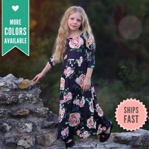 May include: A long, dark blue dress with a floral rose pattern. The dress has a round neckline and three-quarter sleeves. The text "MORE COLORS AVAILABLE" is in a green banner. The dress is worn by a young person.