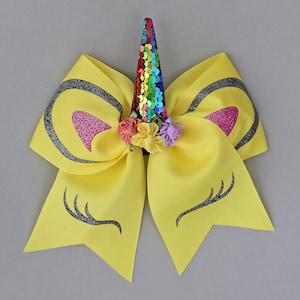 May include: Yellow unicorn hair bow with a rainbow sequin horn, glitter eyelashes, and pink glitter ears. The bow is decorated with small colourful flowers.