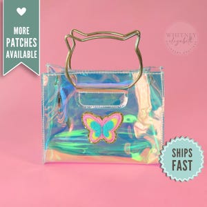 May include: Iridescent clear tote bag with a gold handle and a butterfly patch. The bag has a rectangular shape with a zipper closure. The butterfly patch is in shades of pink, yellow, blue, and green. The bag is on a pink background.