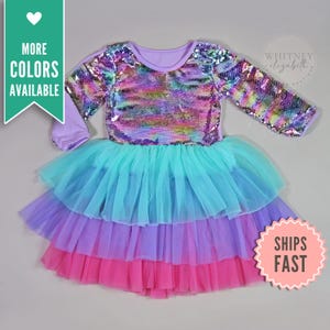 May include: A long-sleeved dress with a sequined bodice and a layered tulle skirt. The bodice features iridescent sequins, while the skirt transitions from turquoise to purple and pink. The dress has a round neckline and is labeled "Whitney Elizabeth Boutique."