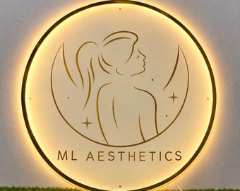 May include: A circular wall sign with a gold-toned frame and warm LED backlighting. The center features a line-art illustration of a person with a ponytail in front of a crescent moon and stars, with the text ML AESTHETICS printed below in gold.