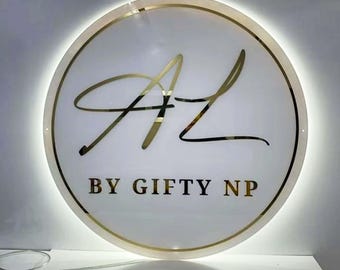 May include: A circular wall sign featuring a white background with a gold metallic border and gold lettering. The sign displays the stylized initials AL in a large script font above the text BY GIFTY NP in a clean, bold, sans-serif typeface.
