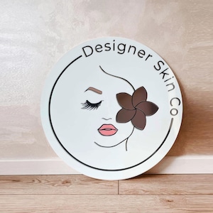May include: White round sign with a black border and the text "Designer Skin Co."  A stylized illustration of a woman's face with closed eyes and a brown flower behind her eye.